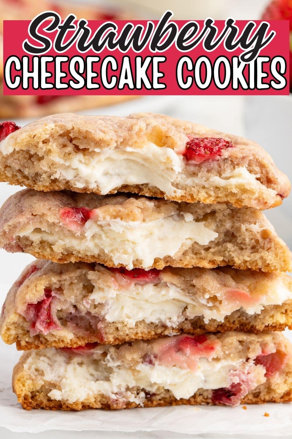A single oversized strawberry cheesecake cookie with a large bite missing, revealing a thick white cream cheese center, resting above a full plate of strawberry cookies piled on a white platter with fresh strawberries visible in the background.