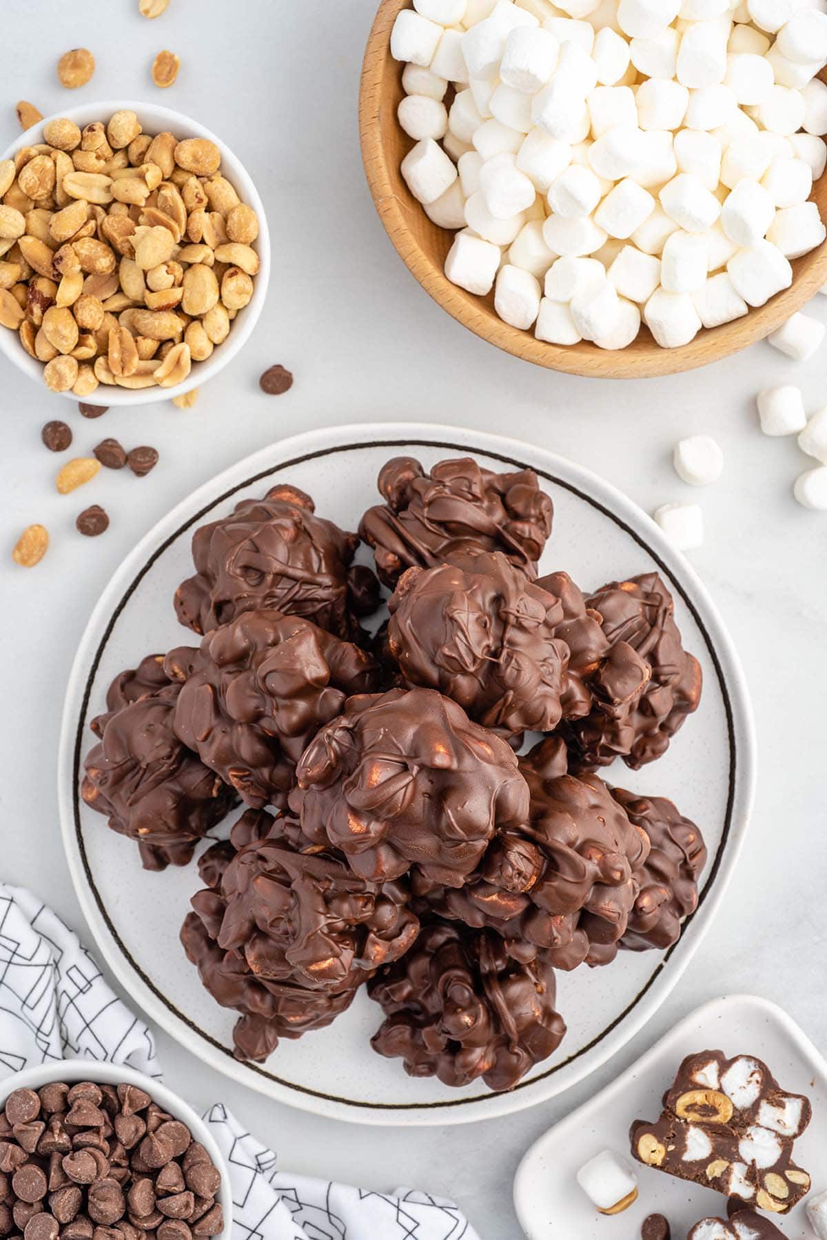 Rocky road candy piled on a plate surrounded by bowls of marshmallows, peanuts, and chocolate chips.