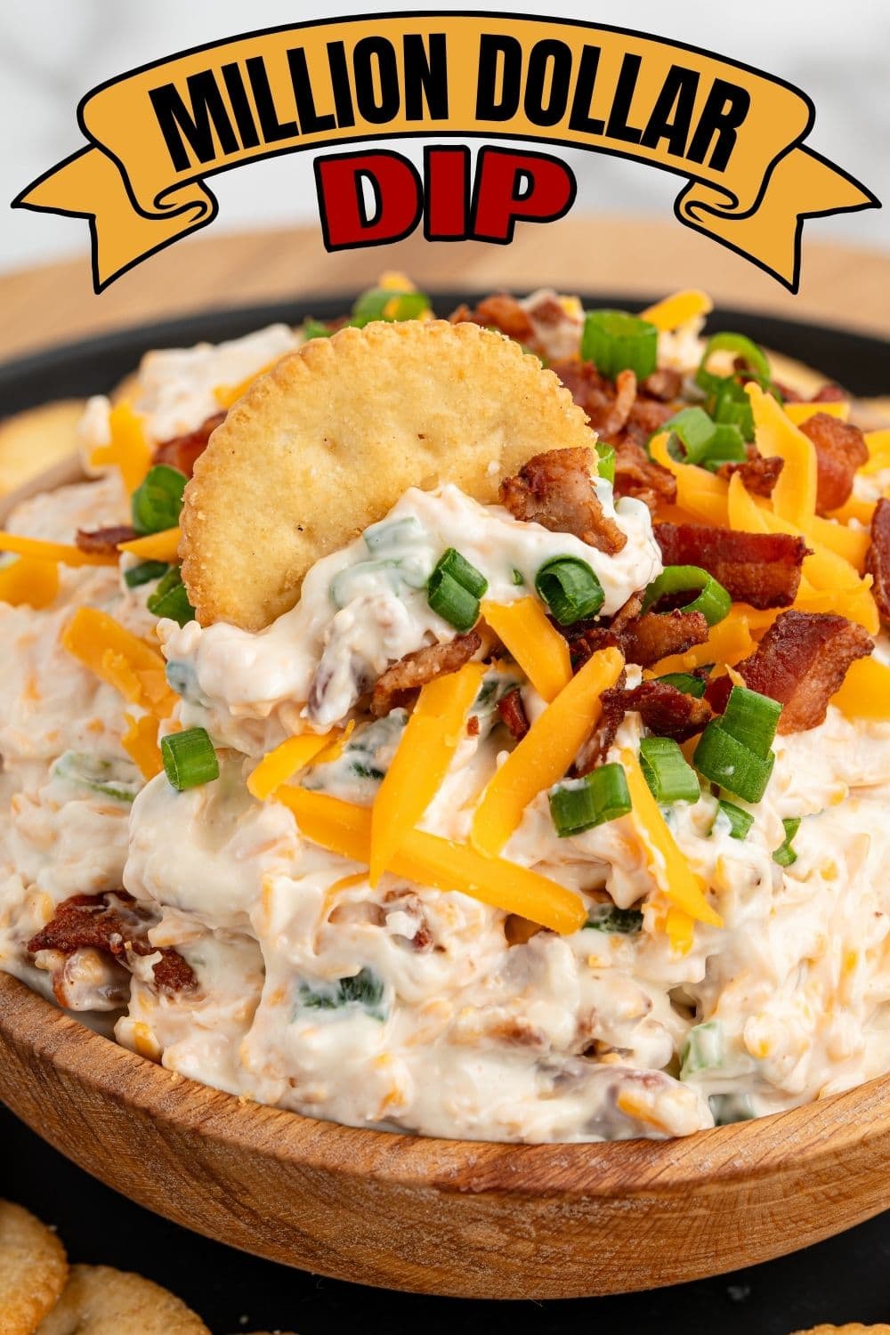 Million Dollar Dip in a wooden bowl topped with shredded cheddar, crumbled bacon, and green onions with a Ritz cracker.