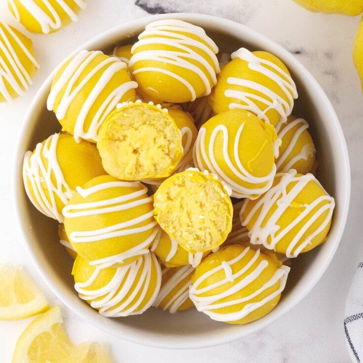 Lemon cheesecake bites piled in a bowl, white chocolate coating with stripes, one bitesize piece cut to expose yellow center, fresh lemons around.