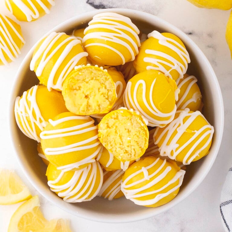 Lemon cheesecake bites piled in a bowl, white chocolate coating with stripes, one bitesize piece cut to expose yellow center, fresh lemons around.