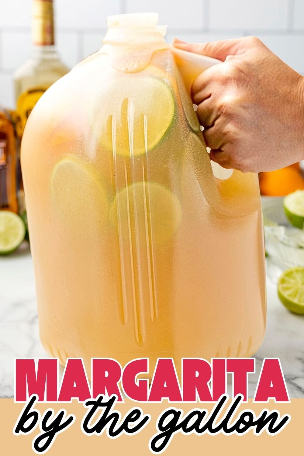 A hand holding margarita by the gallon in a big gallon with a few slices of lime in the background