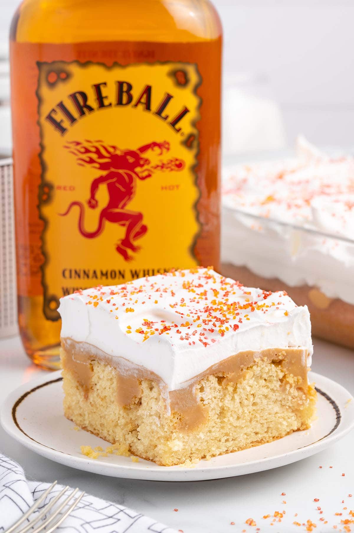 sliced piece of fireball poke cake on a white plate with a bottle of fireball in background.