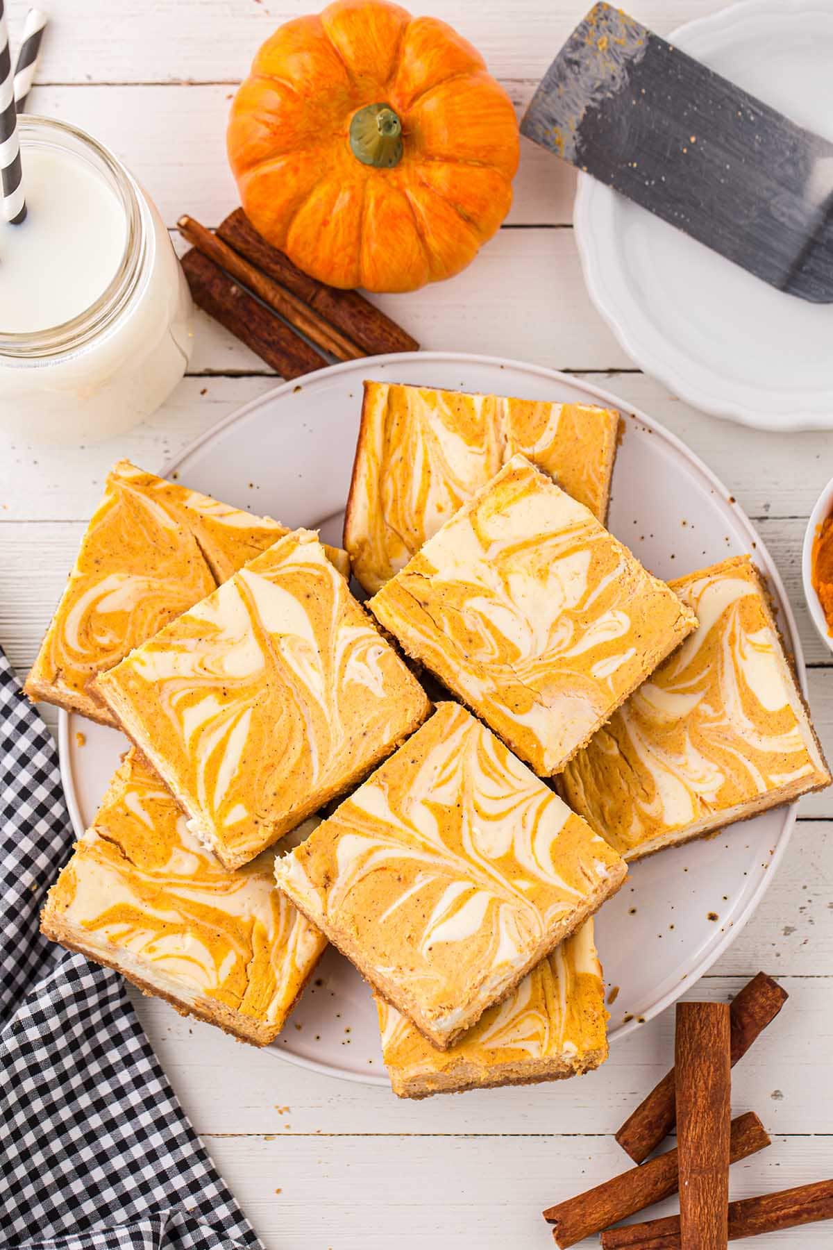 a couple of Pumpkin Cheesecake Bars on a plate. a glass of milk and a pumpkin on the background.