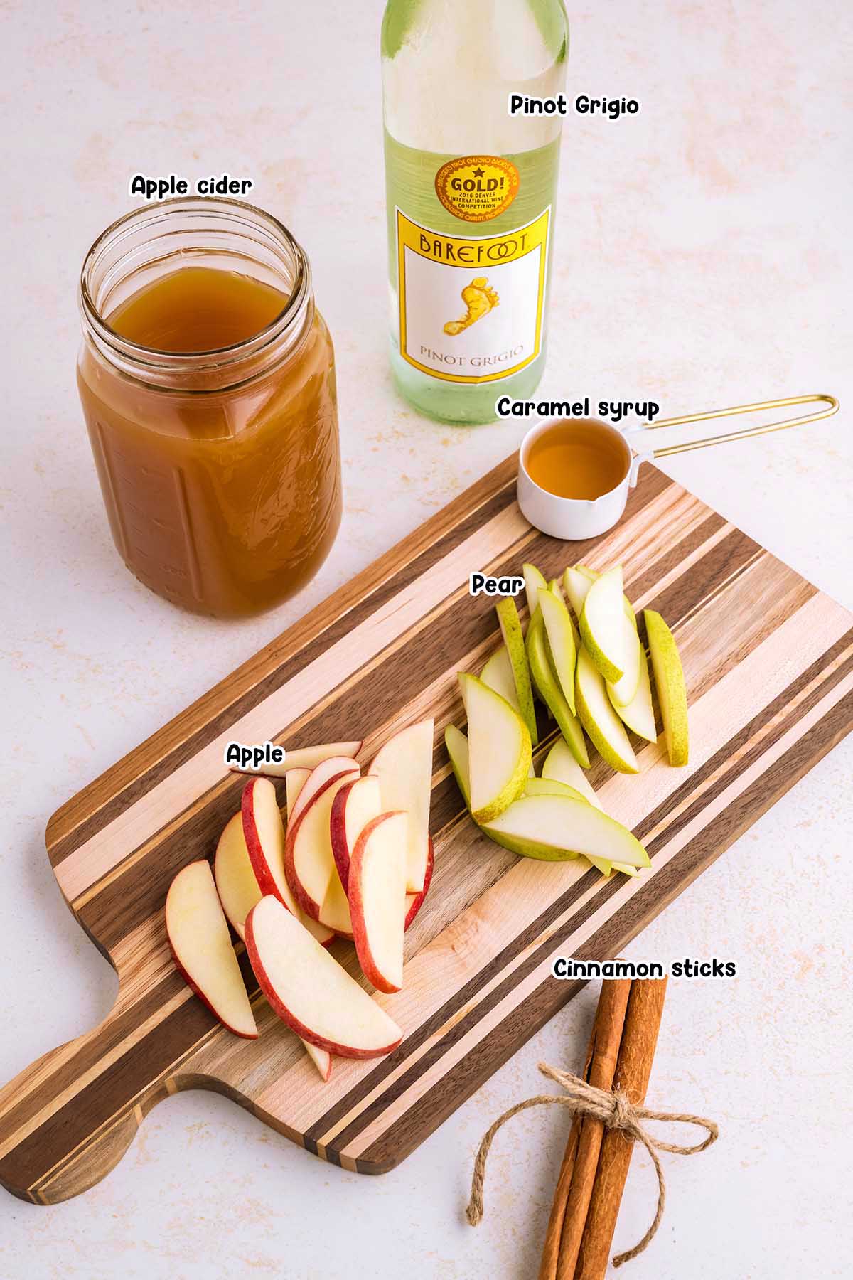 caramel apple sangria ingredients including pinot grigio, apple cider, carmel syrup, sliced apples and pears and cinnamon sticks.