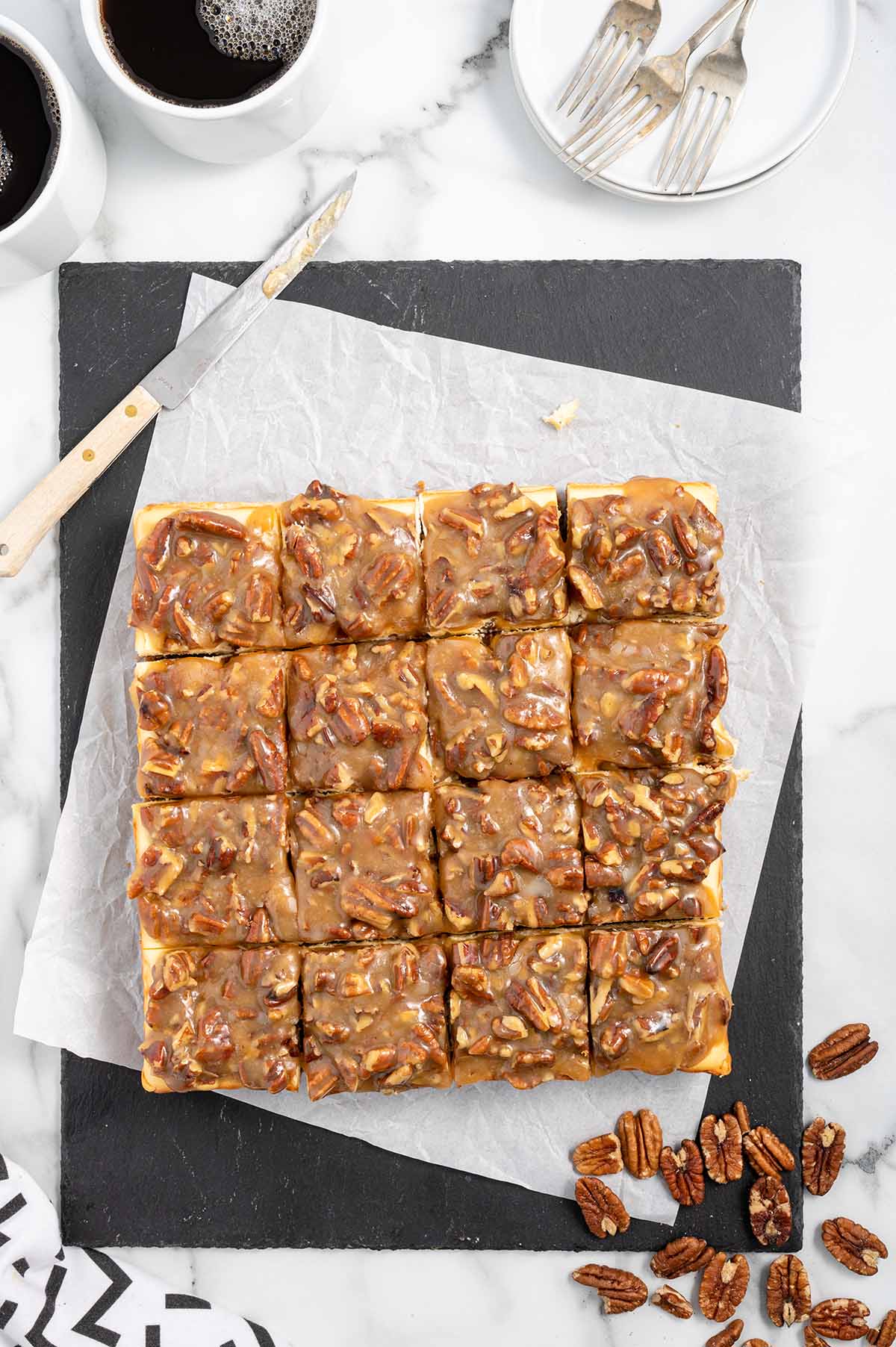 Pecan Pie Cheesecake Bars cut into squares over parchment paper with two cups of coffee on the background.