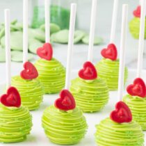 a couple of Grinch Cake Pops on a white table with candy heart and sanding sugar on top.