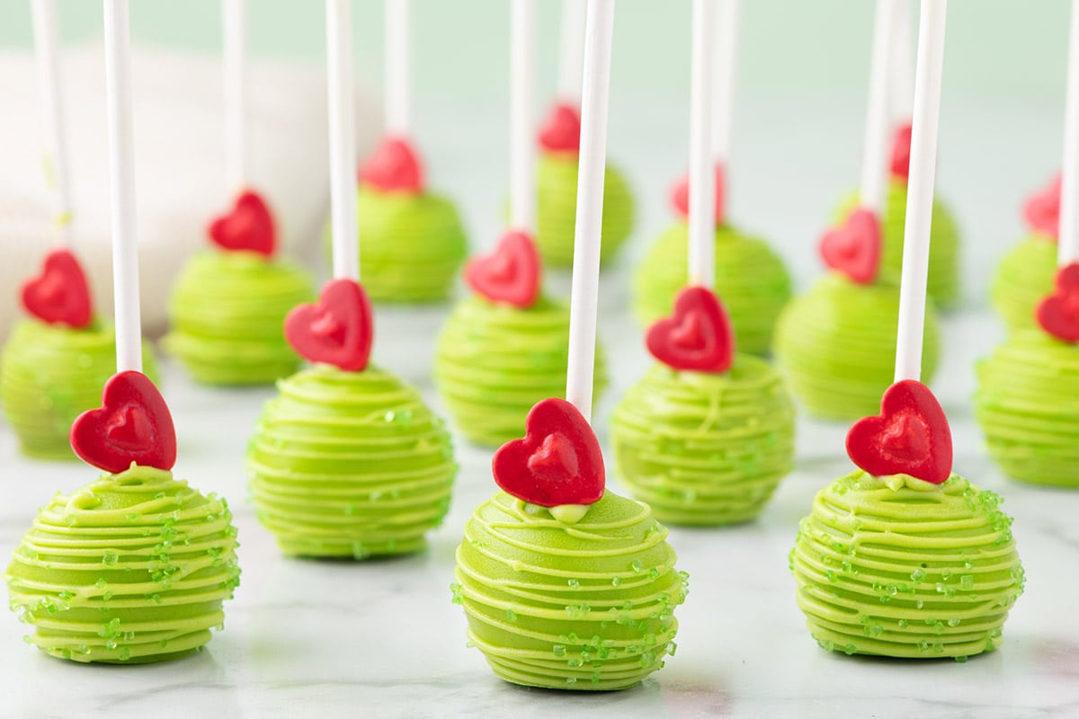 a couple of Grinch Cake Pops on a white table with stick and candy heart.