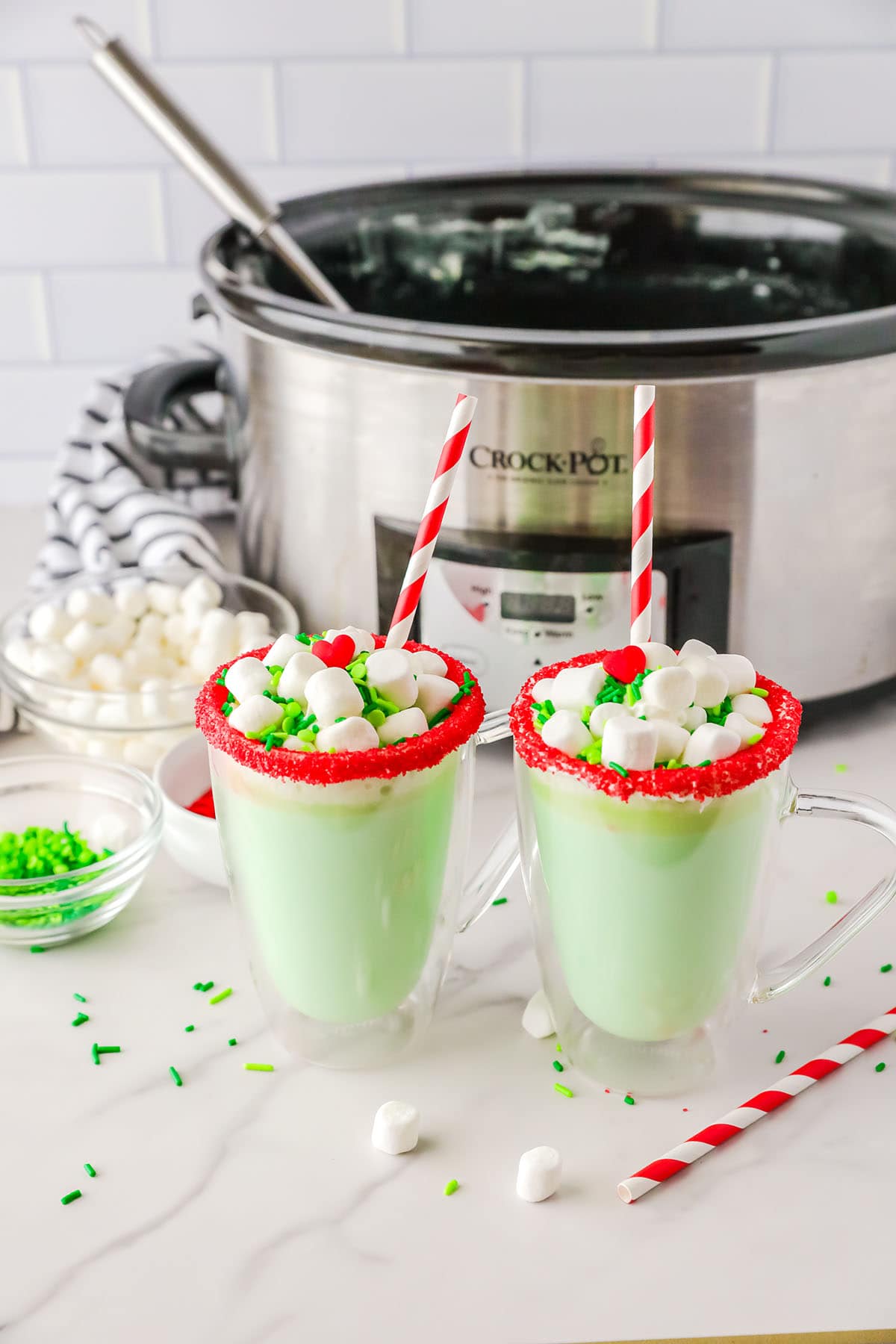 2 mugs of Slow Cooker Grinch Hot Chocolate on a table with marshmallows, sprinkles and candy heart.