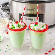 2 glasses of Slow Cooker Grinch Hot Chocolate on a white table with slow cooker on the background.