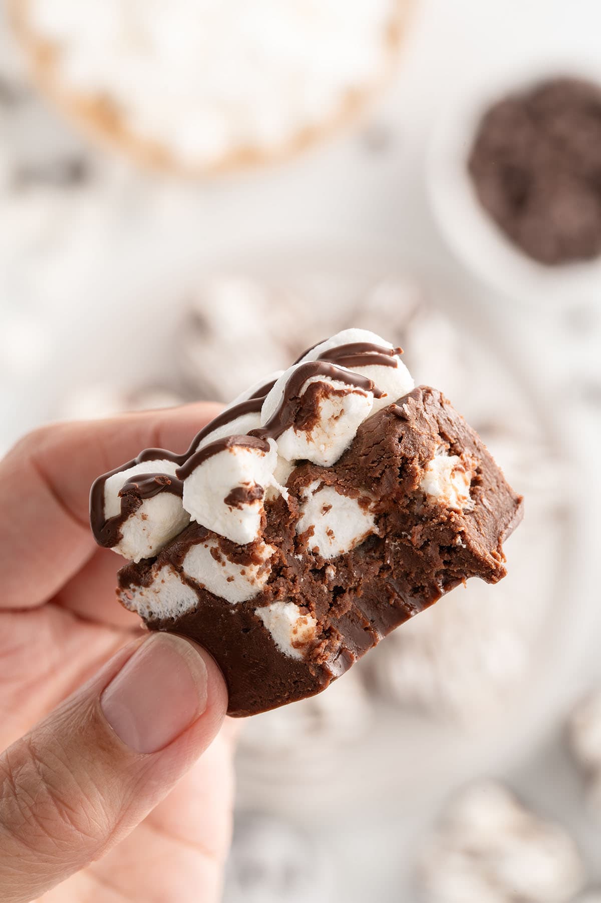 Hand holding a piece of fudge with marshmallows and chocolate drizzle, partially eaten.