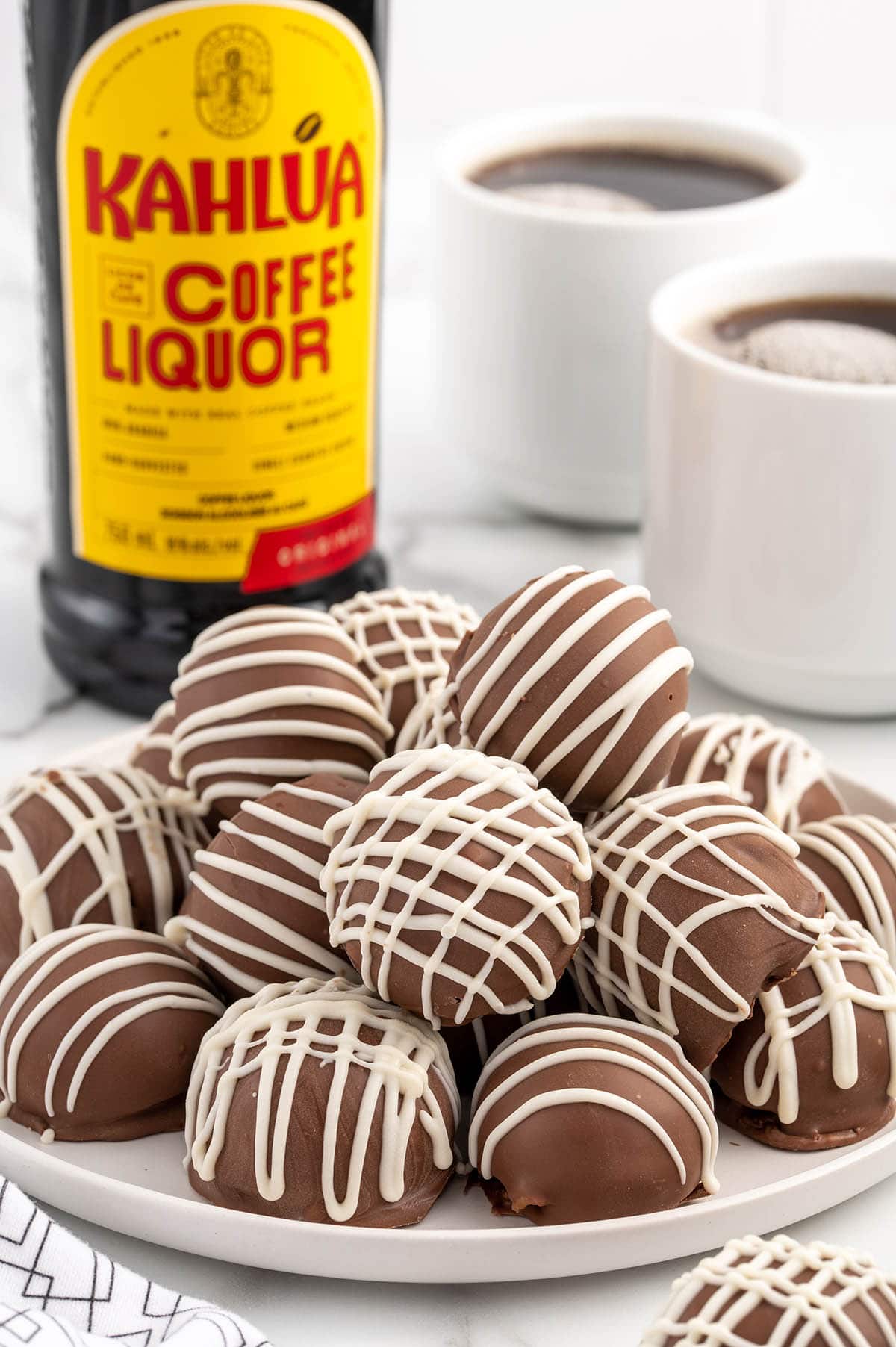 a couple of Kahlua Balls on a white plate with 2 mug of coffee and kahlua bottle on the background.