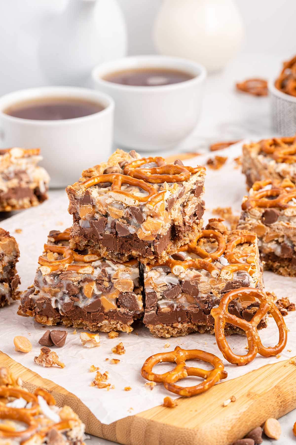 Peanut Butter Pretzel Magic Bars stacked up on a large board with 2 mug of coffee on the background.