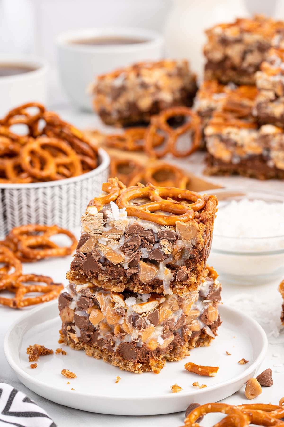 2 bars of Peanut Butter Pretzel Magic Bars on a white plate and pretzels in a bowl on the background.
