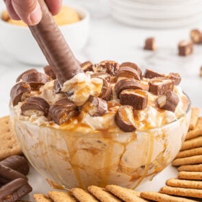 dipping twix bars into Twix Dip in a bowl with some crackers surrounding it.