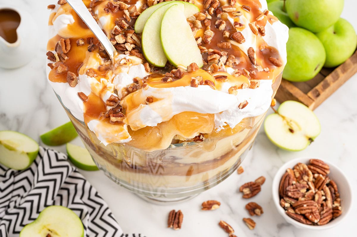 Caramel Apple Trifle on a white table with chopped pecan for toppings and a couple apples on the background.