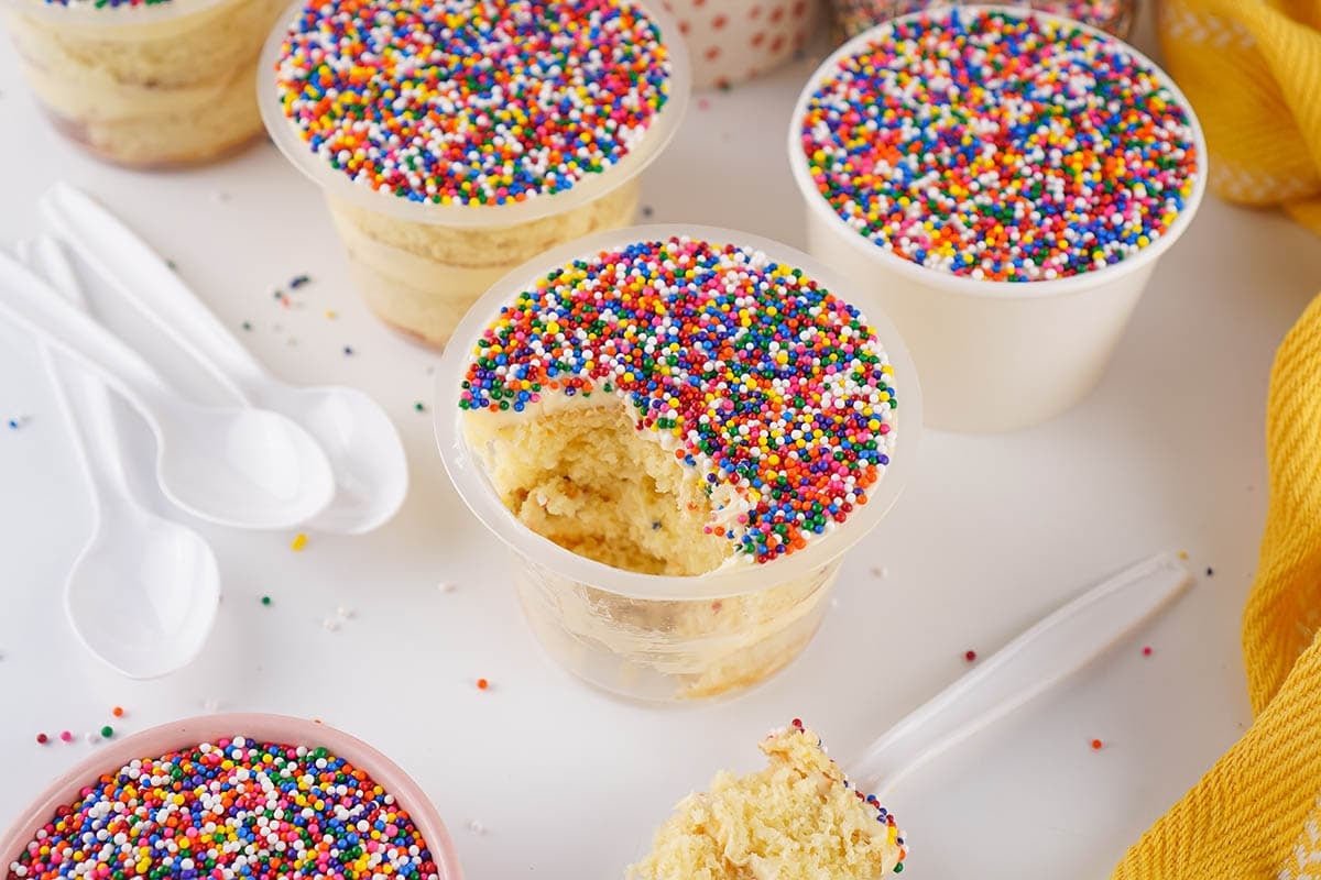Several vanilla cake cups topped with rainbow nonpareil sprinkles, with one cup scooped open showing the cake inside.