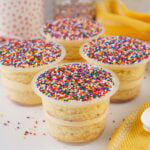 Four layered vanilla Dot Cakes in clear plastic cups topped with rainbow nonpareil sprinkles.