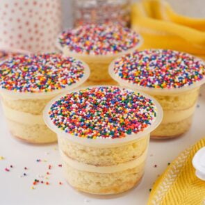 Four layered vanilla Dot Cakes in clear plastic cups topped with rainbow nonpareil sprinkles.
