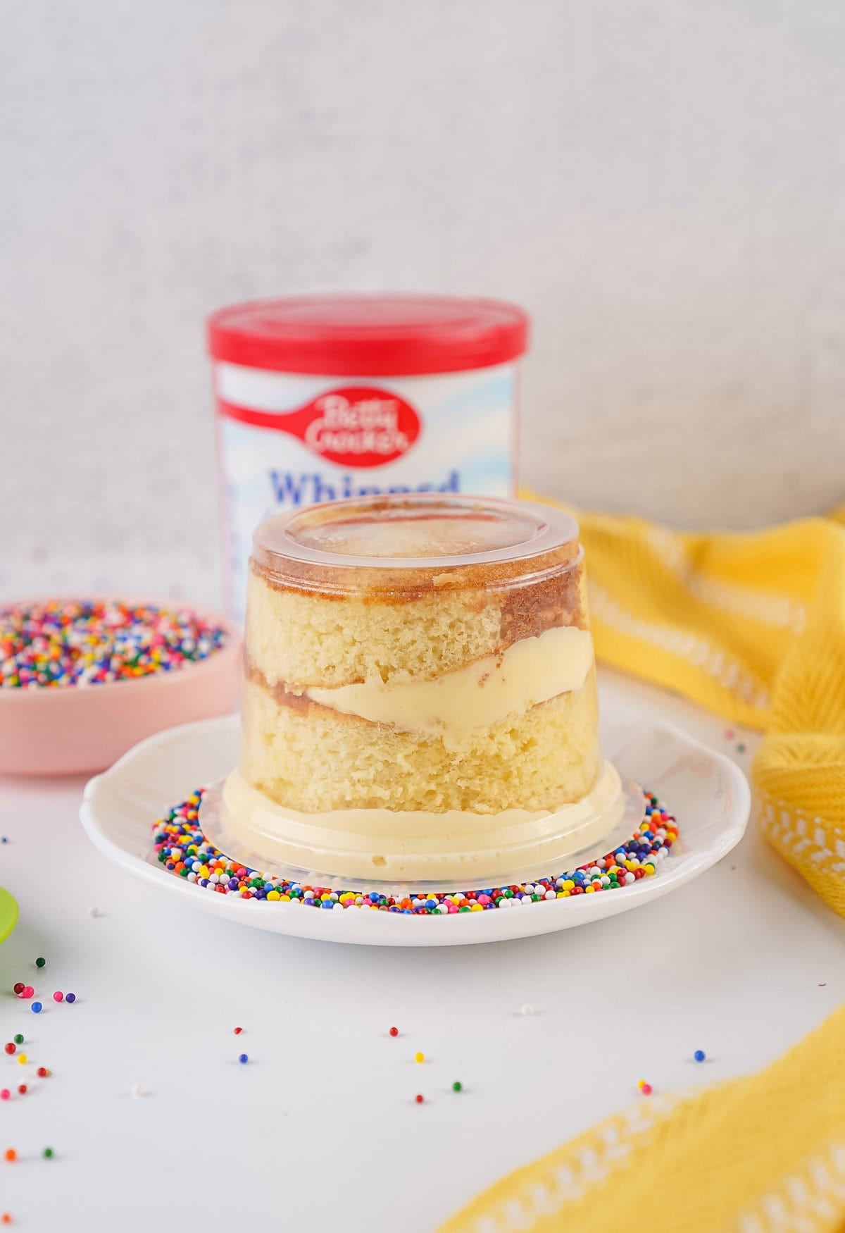 Layered vanilla cake and frosting in a clear dessert cup with rainbow sprinkles nearby