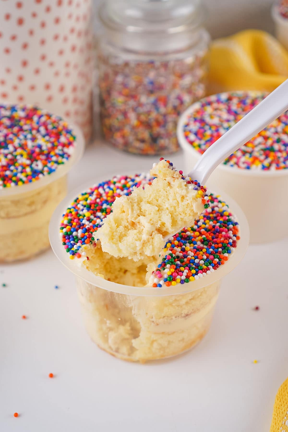 A spoonful of vanilla cake being lifted from a plastic cup topped with colorful rainbow nonpareil sprinkles.