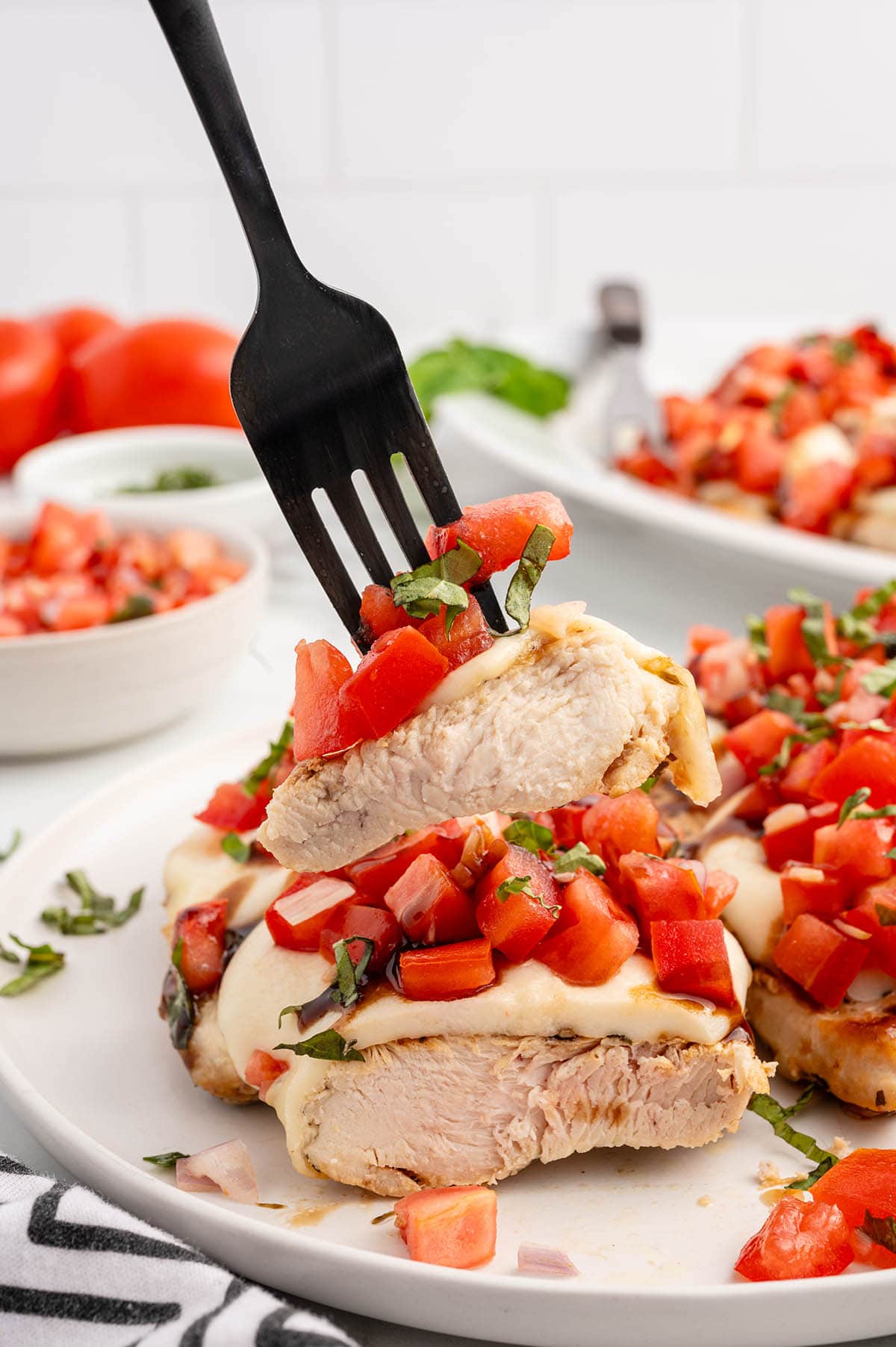 Fork holding a slice of bruschetta chicken with tomatoes and basil on a white plate.