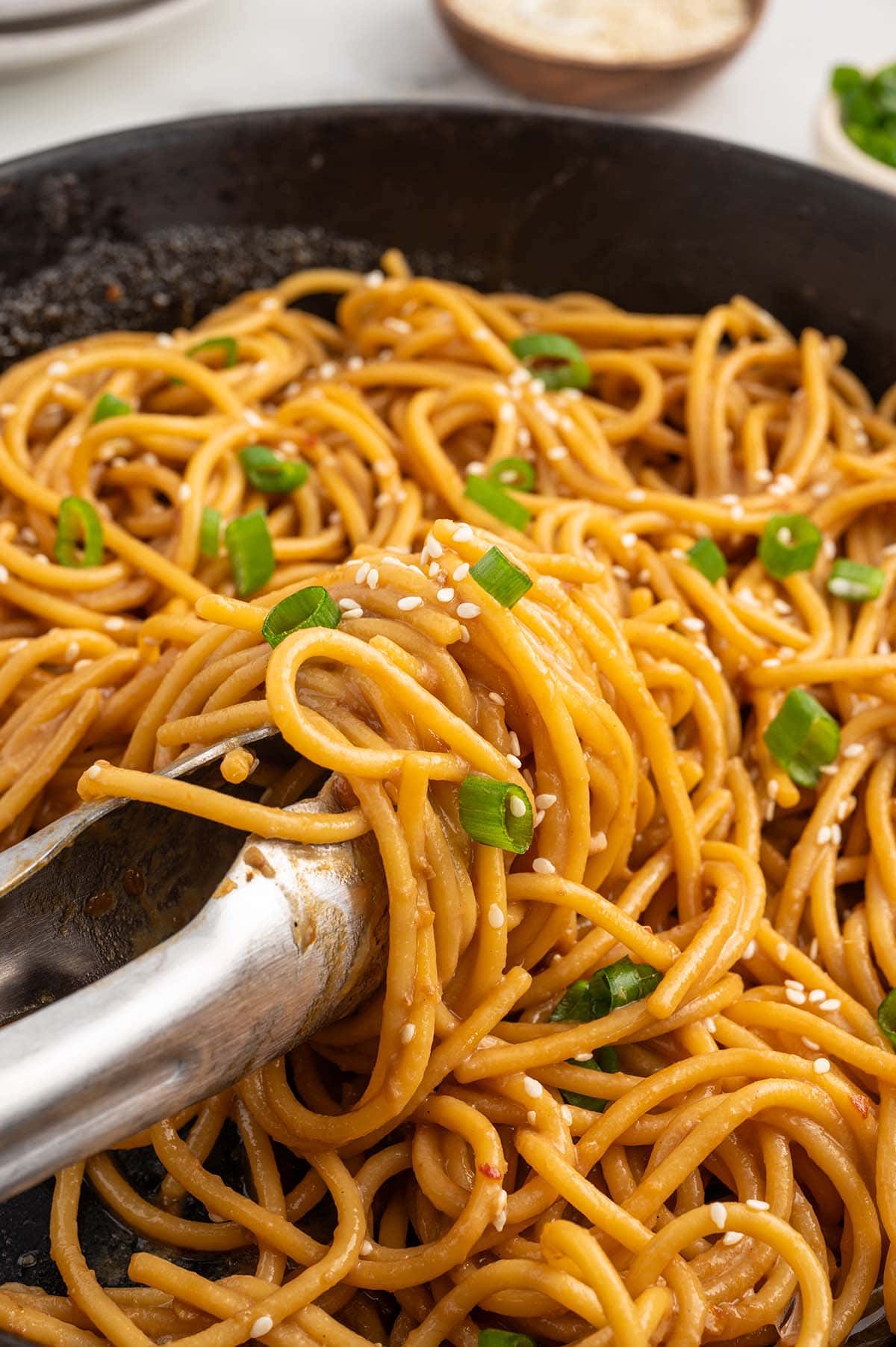 using tongs to take Garlic Sesame Noodles from the skillet.
