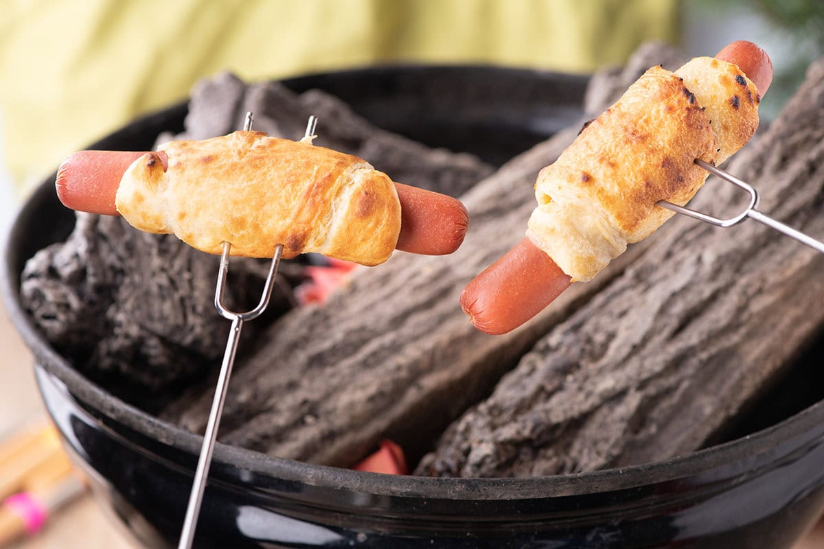 cooking Campfire Crescent Roll Hot Dogs on a skewer.