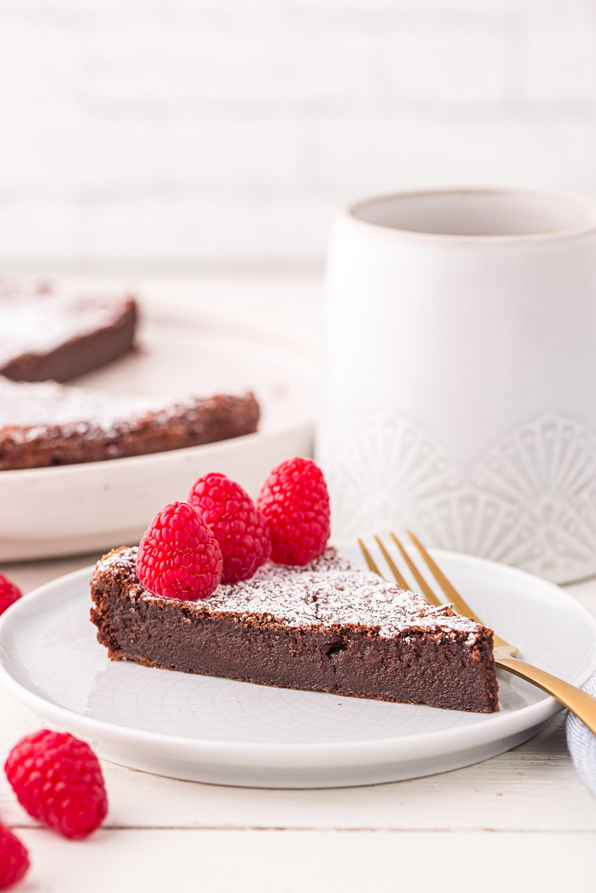 a slice of Flourless Chocolate Torte on a white plate with powdered sugar and raspberries.