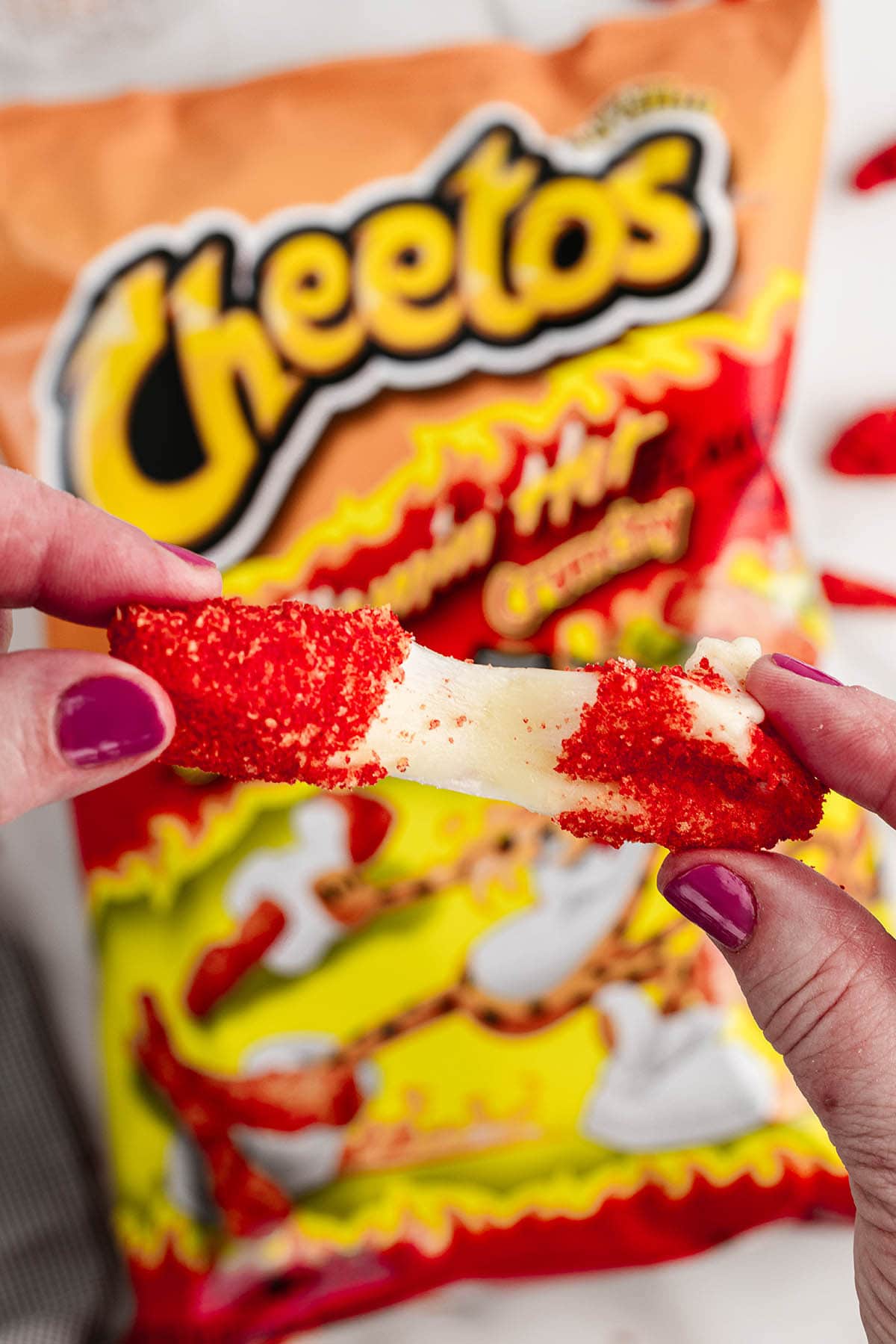 hand stretching out a piece of Hot Cheeto Cheese Sticks with a bag of cheetos at the back.