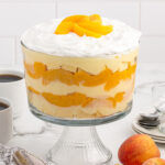 Peaches and Cream Trifle in a trifle bowl and some peaches on the table.