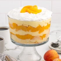 Peaches and Cream Trifle in a trifle bowl and some peaches on the table.