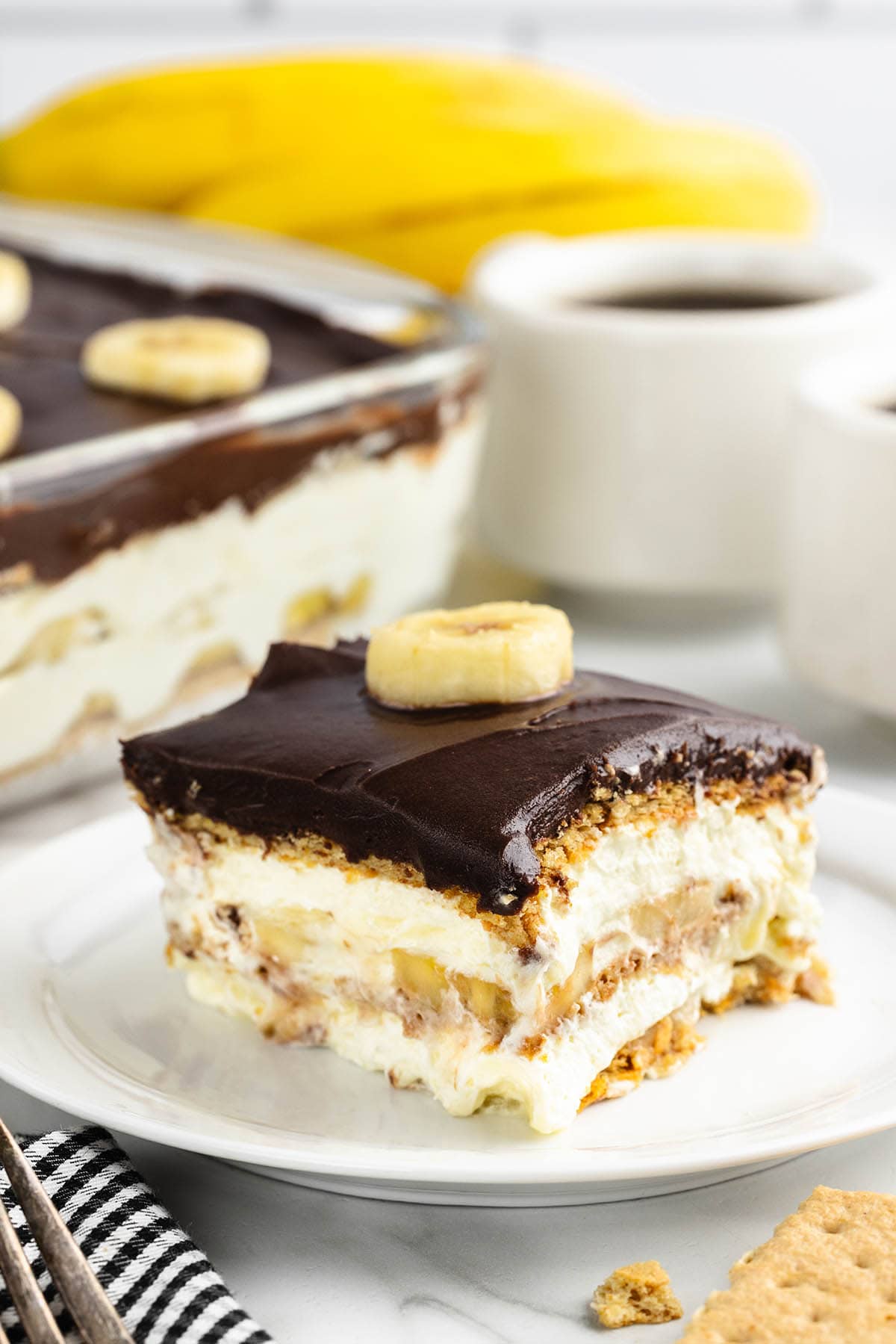 a slice of Banana Eclair Cake on a white plate topped with a layer of chocolate frosting and a banana slice.