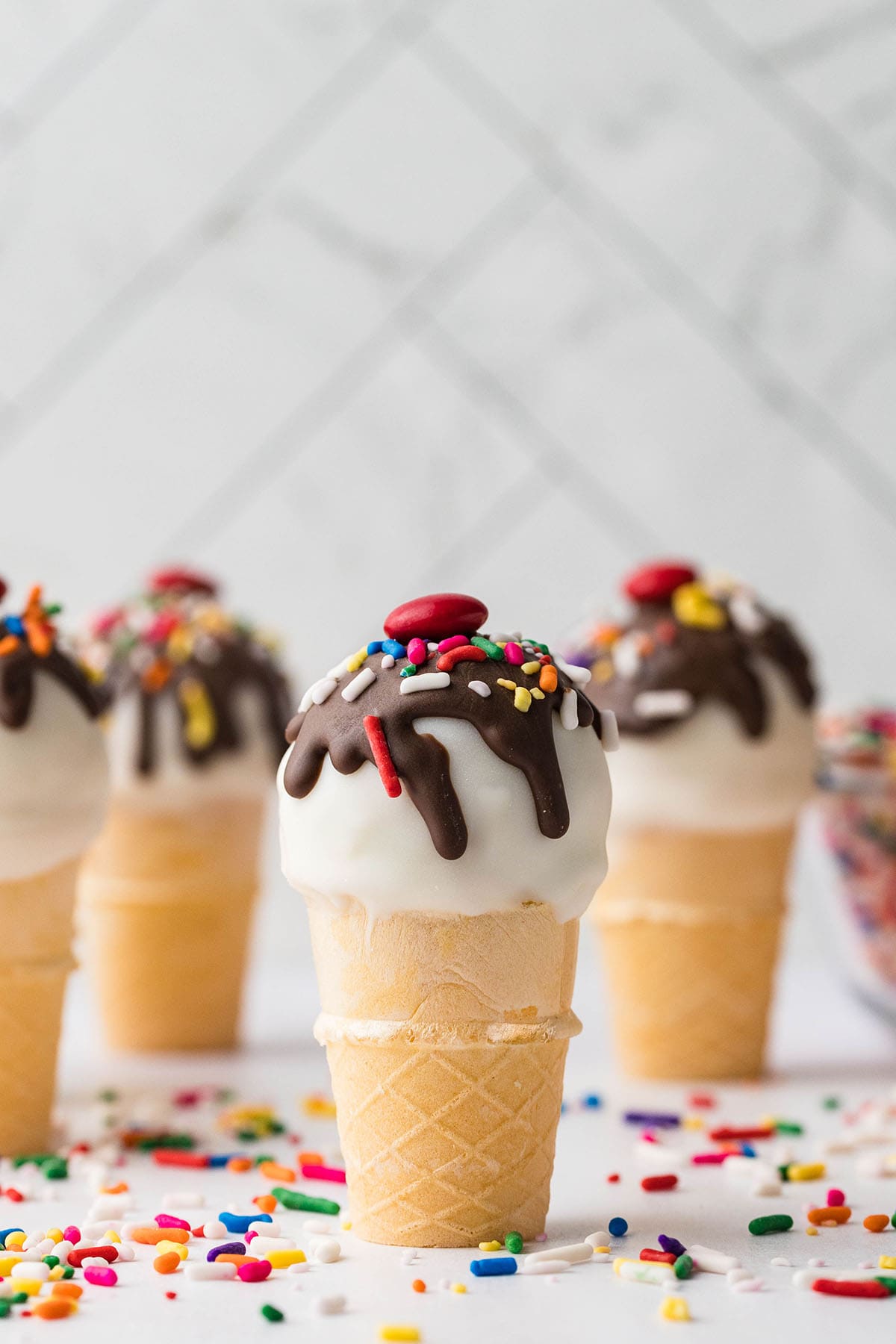 a couple of Cake Pops Ice Cream Cones on the table with sprinkles and M&M's on top.