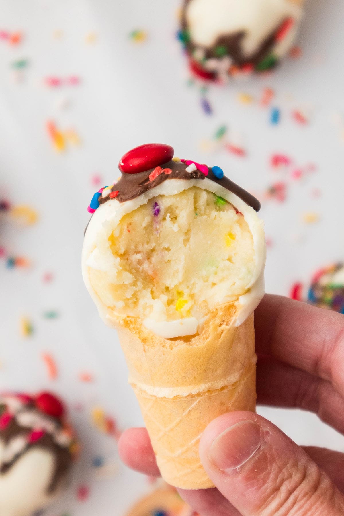 hand holding a Cake Pops Ice Cream Cone partially eaten with sprinkles in the background.