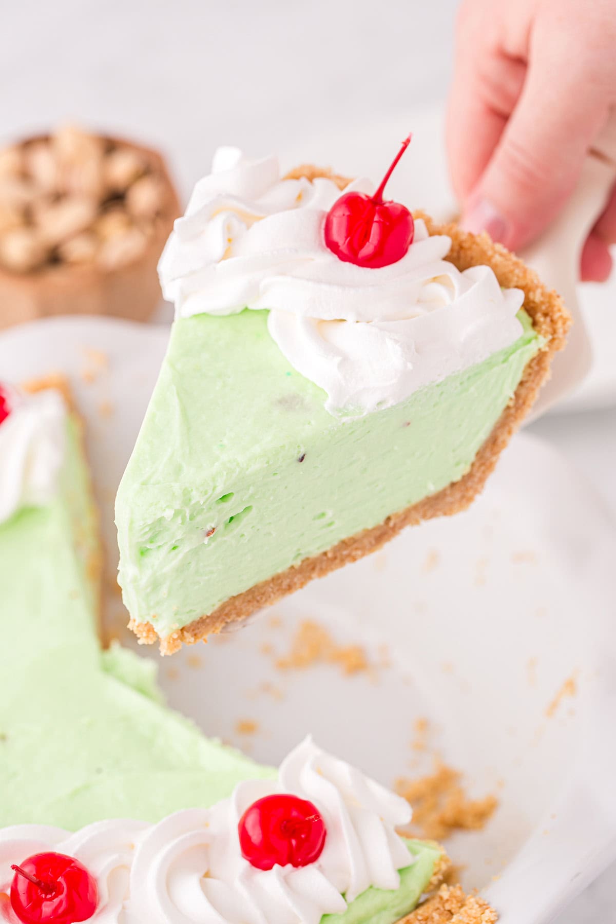 a slice of Pistachio Pie with whipped cream and a cherry on top, being lifted from the pie pan. 