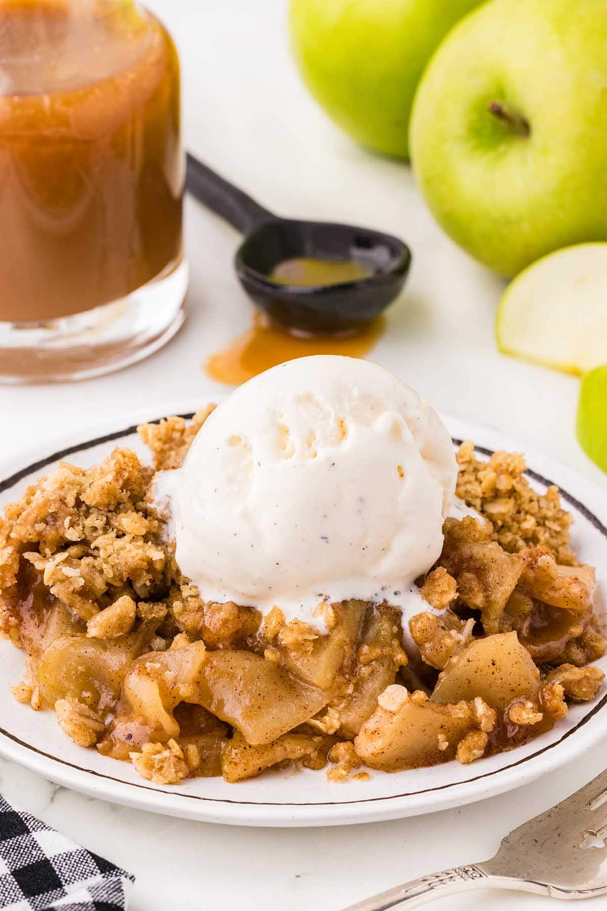 a bowl of Apple Crisp topped with a scoop of ice cream and caramel sauce in the background.