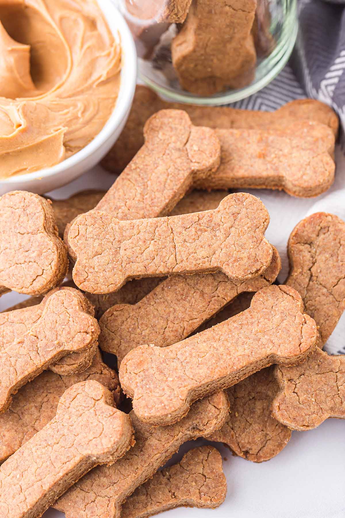 Peanut Butter Dog Treats scattered on a white table and a bowl of peanut butter on the side.