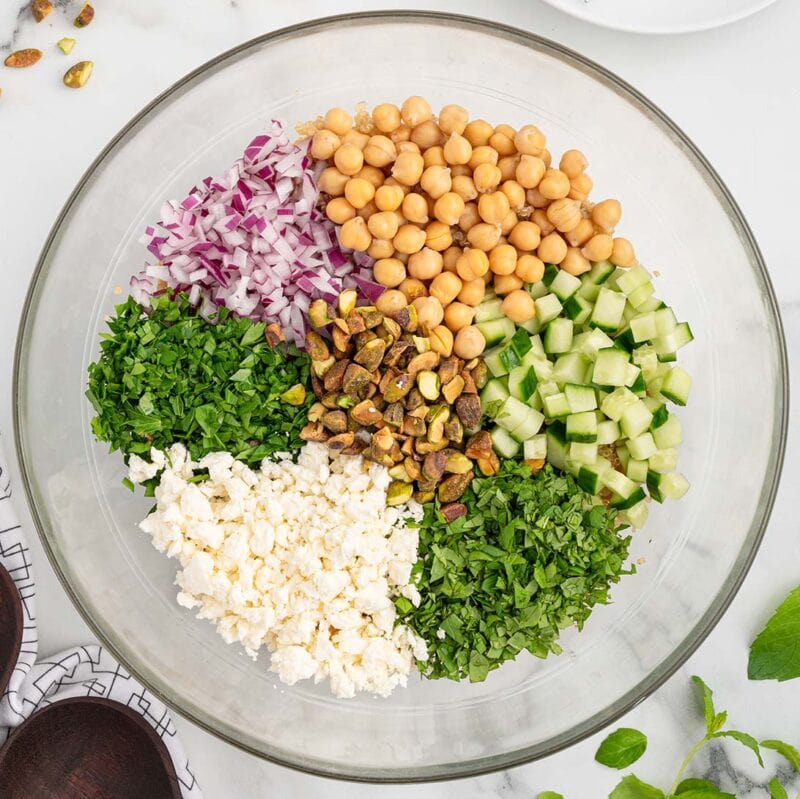 a bowl filled with chickpeas, cucumber, red onion, pistachio nuts, feta cheese crumbles, fresh parsley and fresh mint.