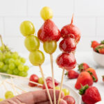 a hand holding three skewers of Tanghulu with glazed grapes and strawberries.