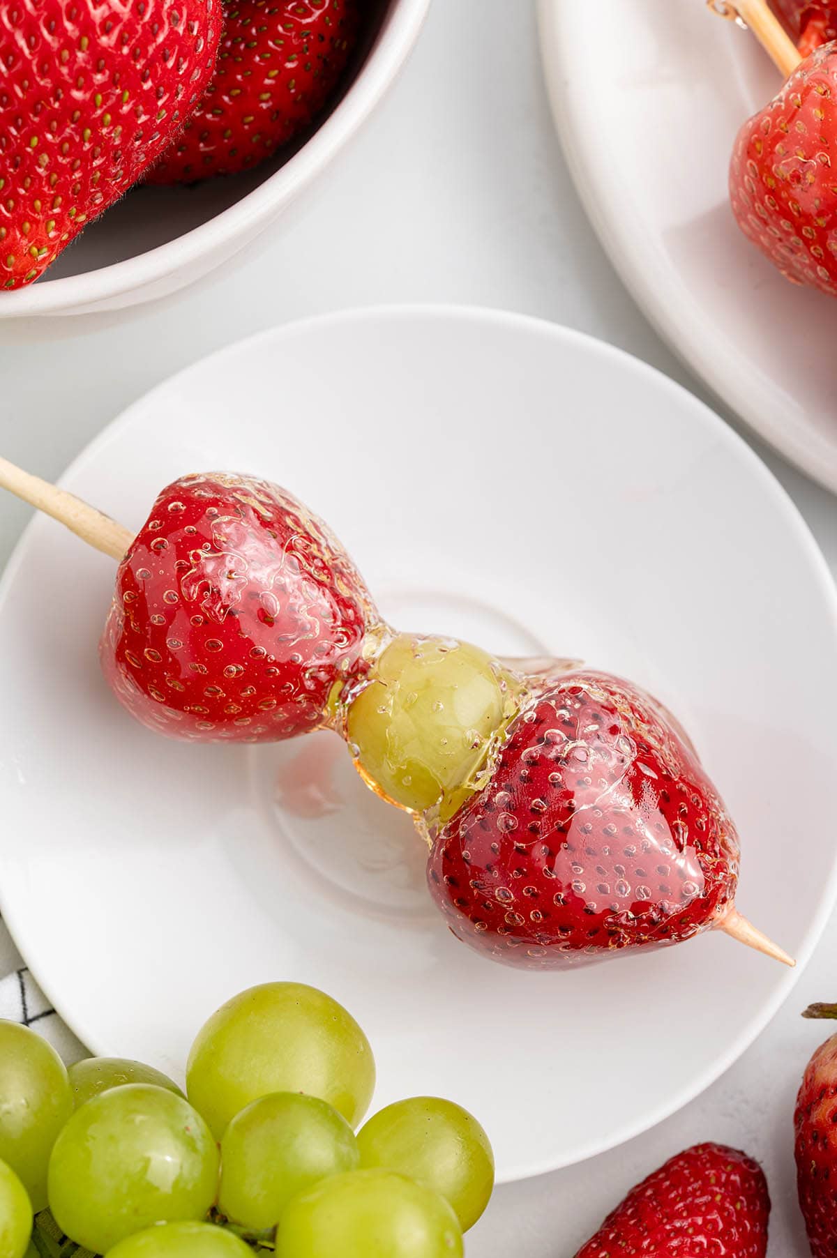 a skewer of Tanghulu with two strawberries and a green grape coated in a glossy, hard candy shell.