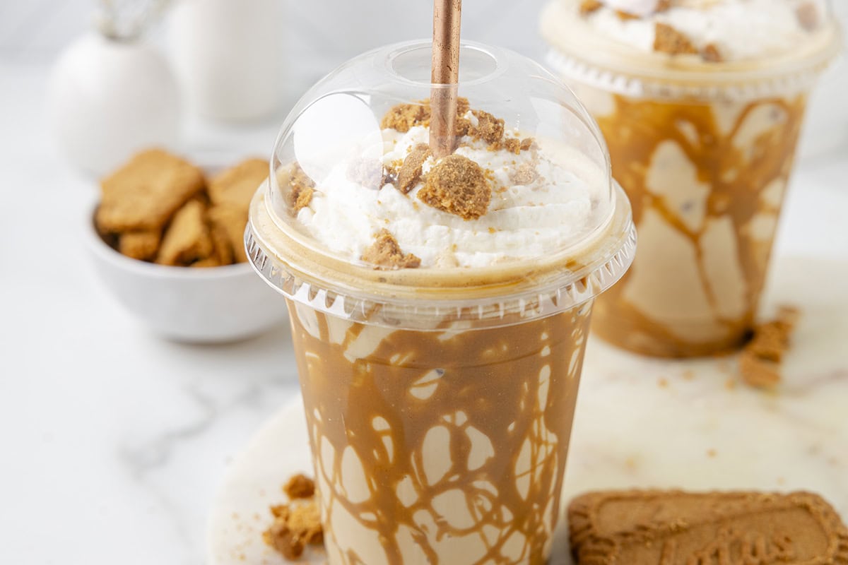 Iced Cookie Butter Latte in a plastic cup with whipped cream and crumbled cookies in the background is another cup and a bowl of cookies.