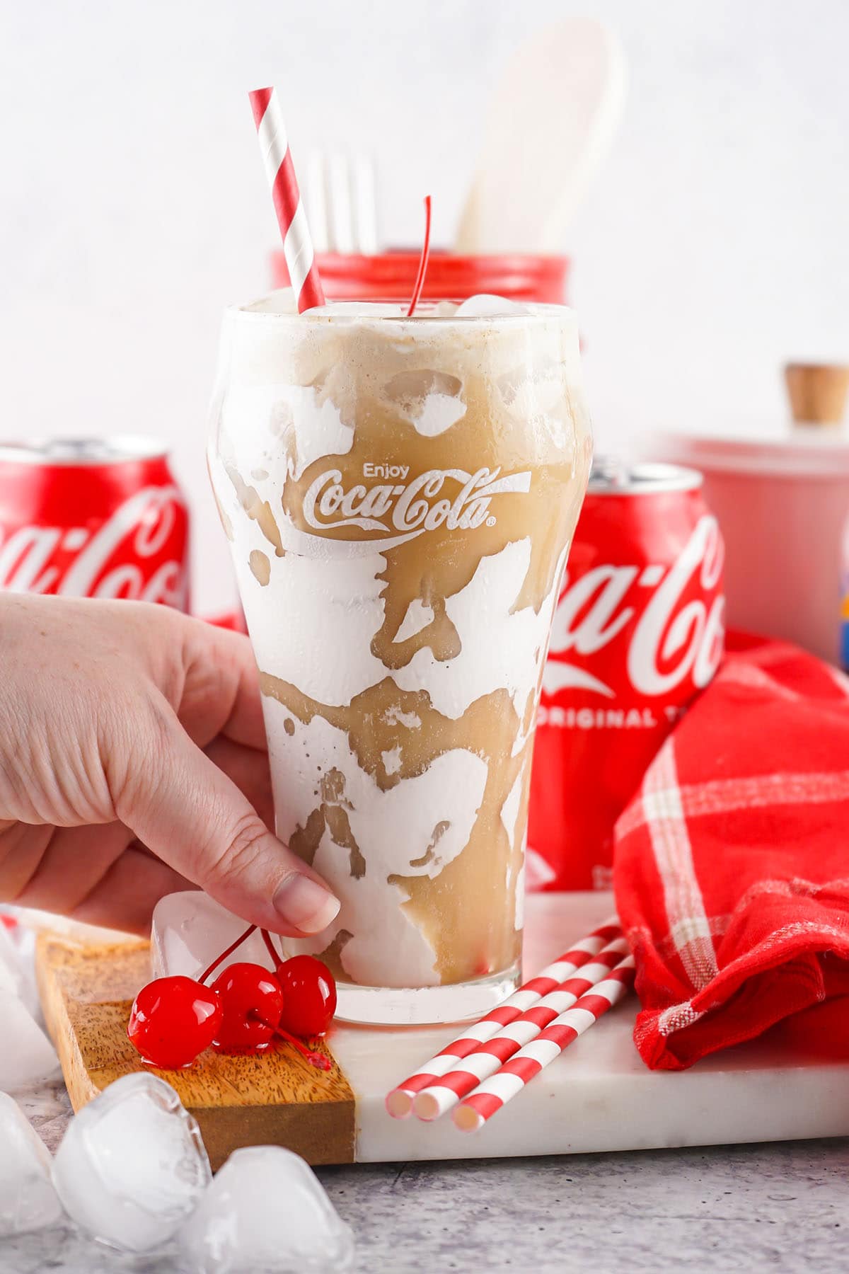 hand holding a glass of Fluffy Coke with extra cherries and straws on the table.