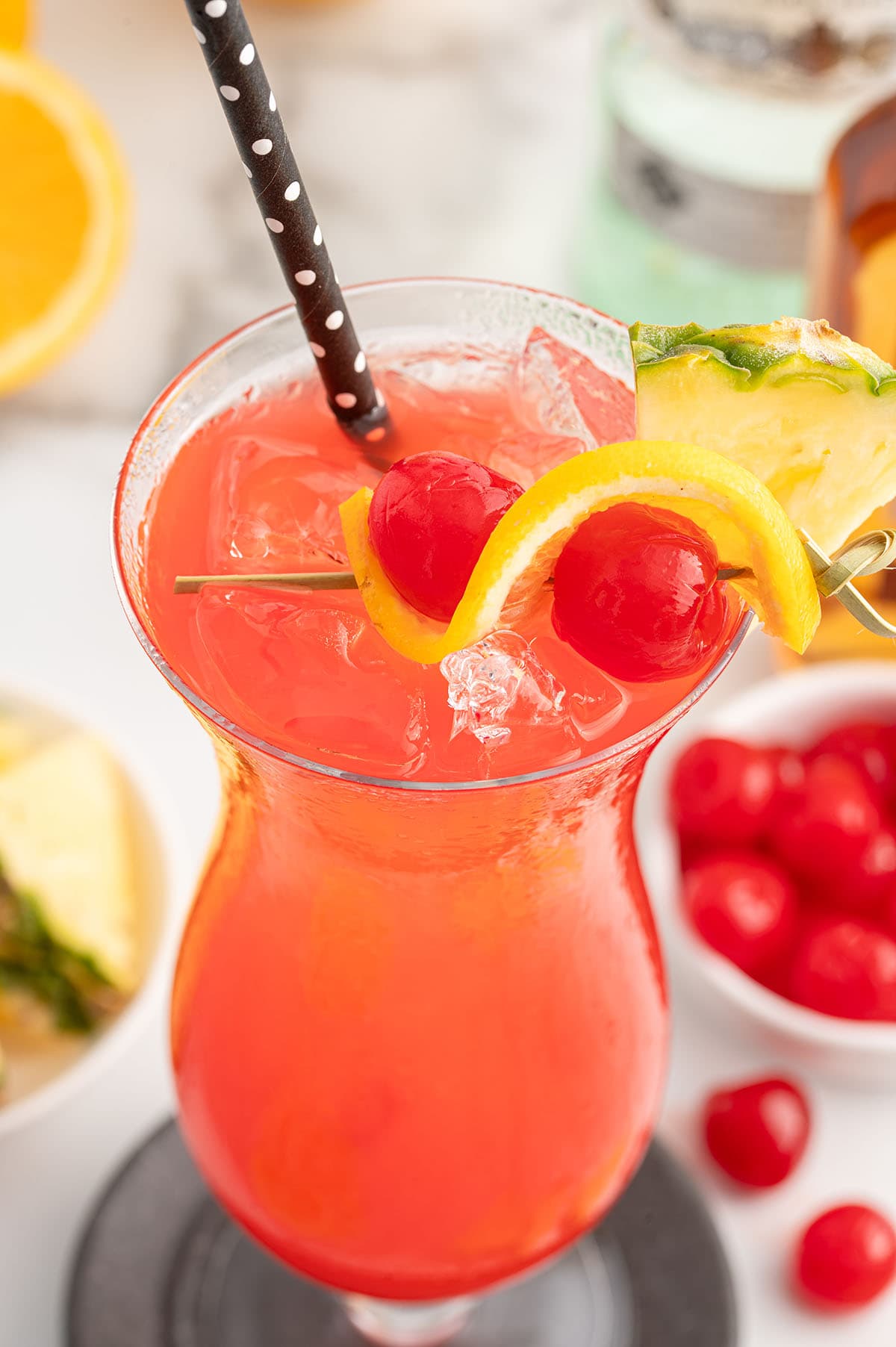 Rum Punch in a hurricane glass garnished with lemon, pineapple slice and maraschino cherries with a black straw.