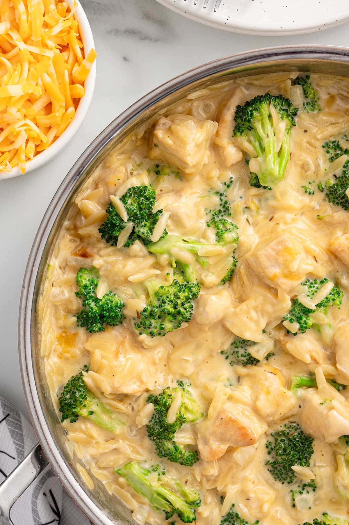 a cheesy chicken orzo with broccoli florets in a large skillet and a bowl of shredded cheese.