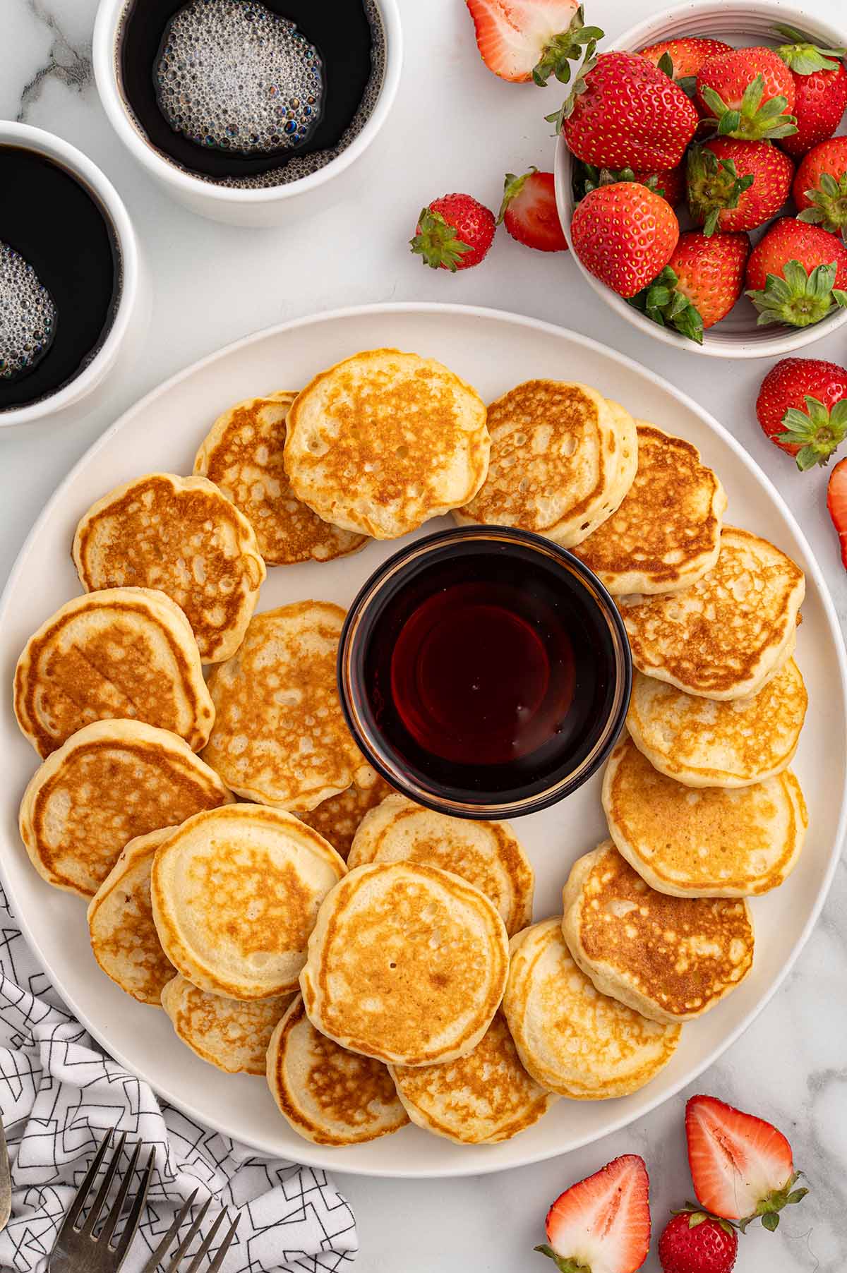 a couple of mini pancakes on a white plate with a bowl of syrup in the middle. 