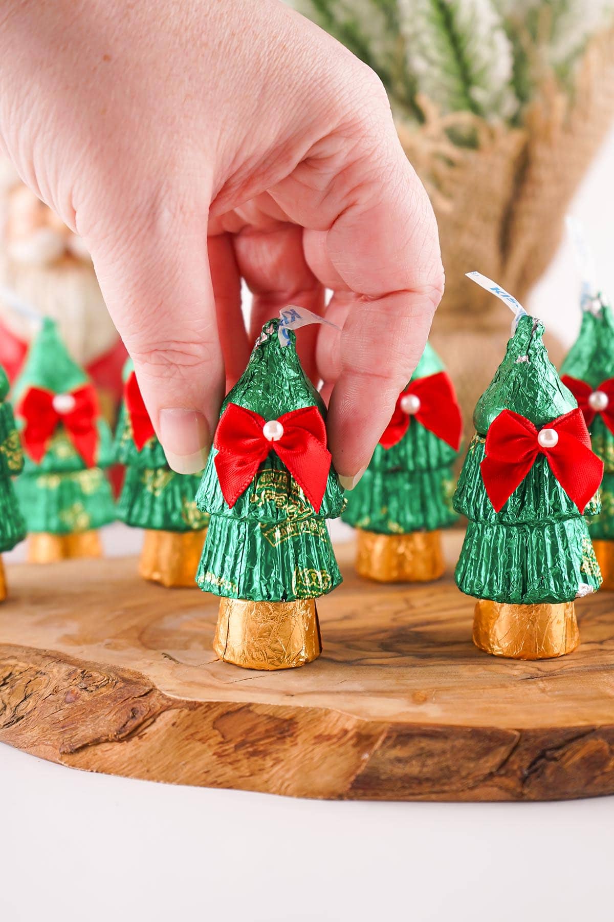 A hand arranging a candy Christmas tree with green foil, red bow, and pearl on a wooden surface.