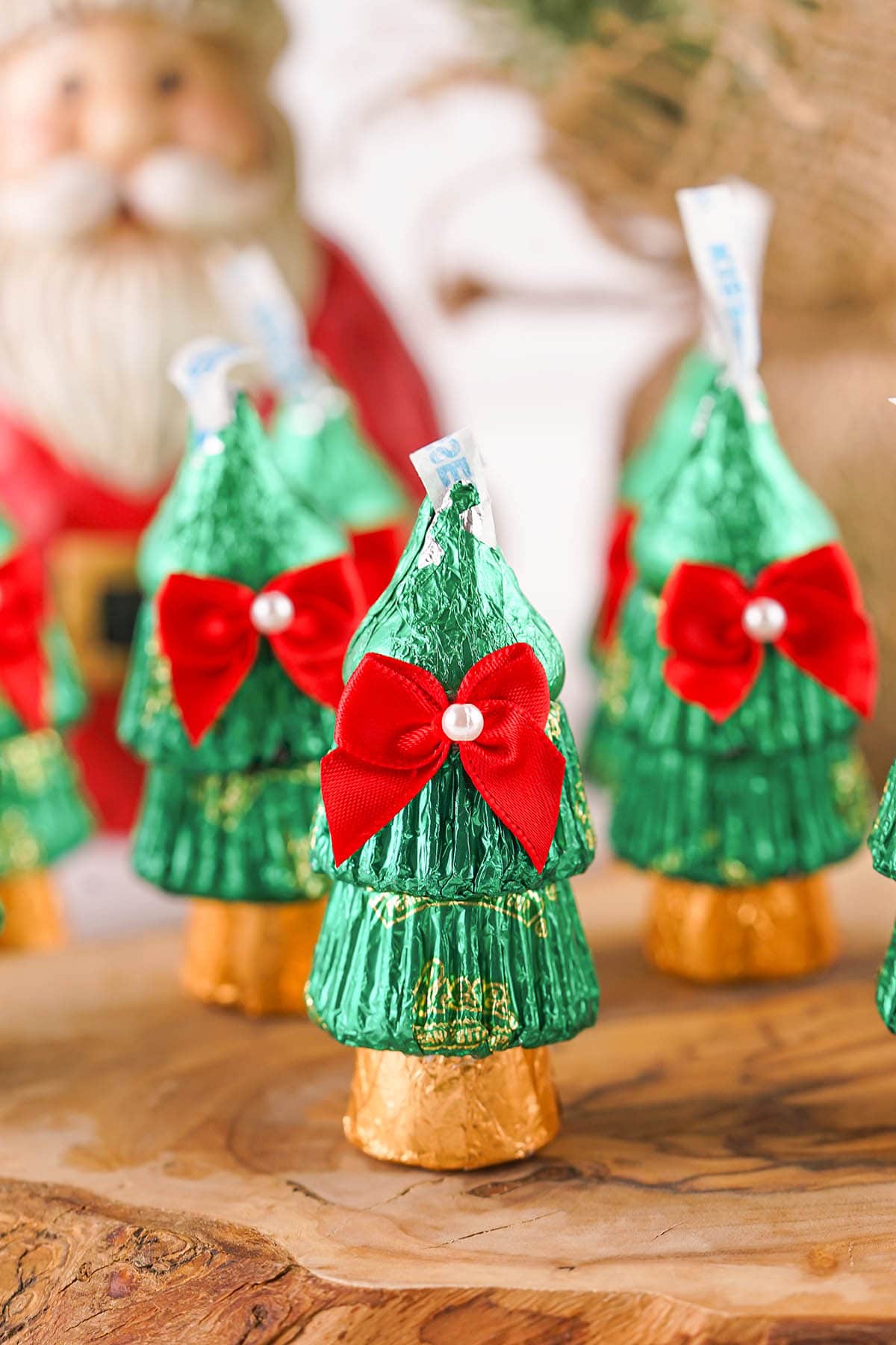 Candy Christmas trees with green foil, red bows, and pearl accents, on a wooden surface with Santa in the background.