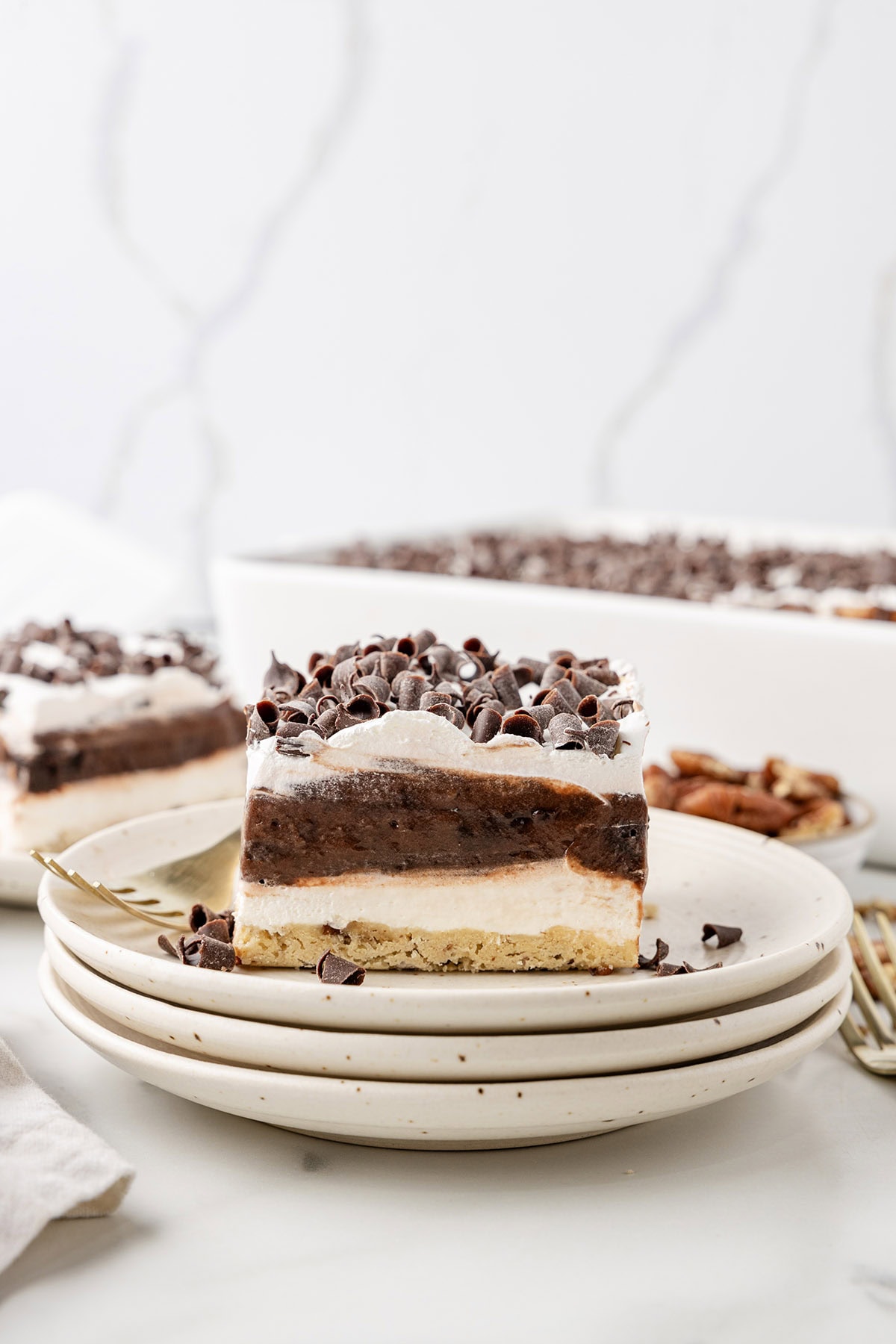 A slice of layered chocolate delight on stacked plates, topped with chocolate curls, with the full tray in the background.