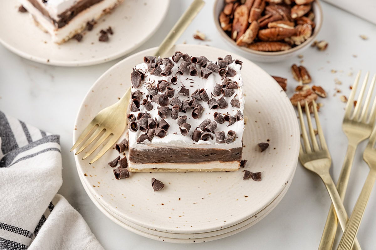 Chocolate delight topped with chocolate curls, served on a plate with golden forks and pecans in the background.